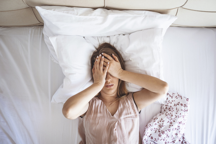 Young beautiful Caucasian woman on bed having headache / insomnia / migraine / stress. Young woman covering her face with hands. Desperate girl suffering insomnia trying to sleep