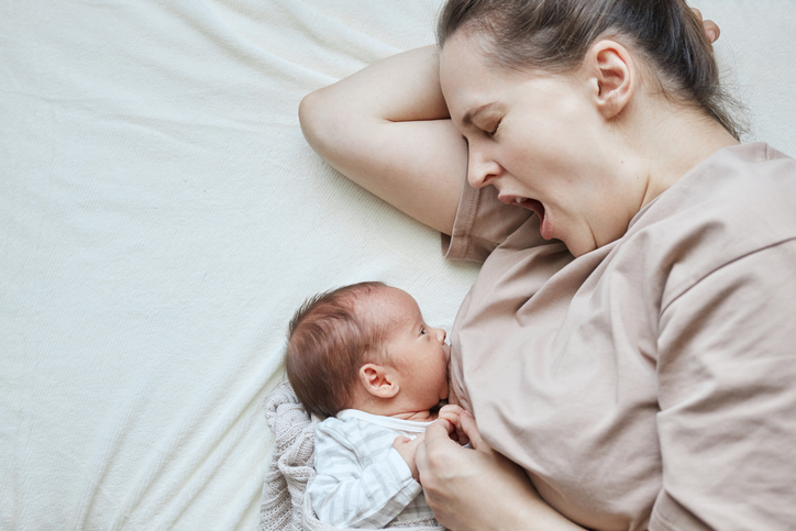 Home milk feeding. Home breastfeeding routine. Sleepy yawning mother breastfeeding her baby while lying on bed being tired after sleepless night