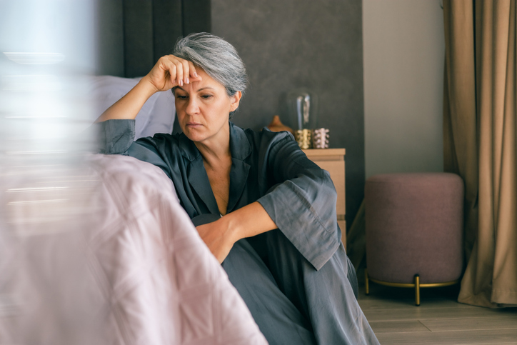 A mature woman with gray hair sits on the bedroom floor, looking exhausted and deep in thought, reflecting midlife stress, emotional burnout, or menopause challenges.